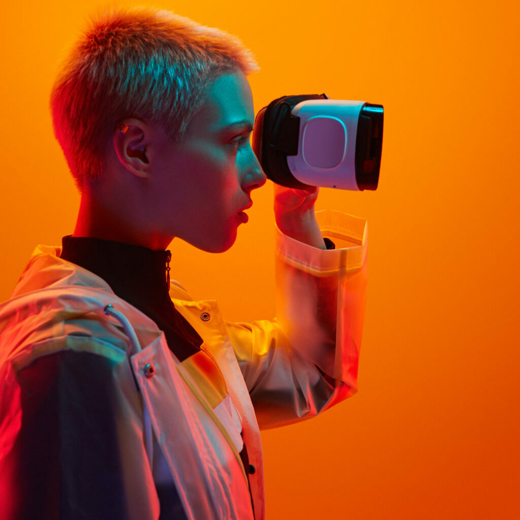 Side view of emotionless female experiencing metaverse while using virtual reality helmet on orange background in studio with neon illumination